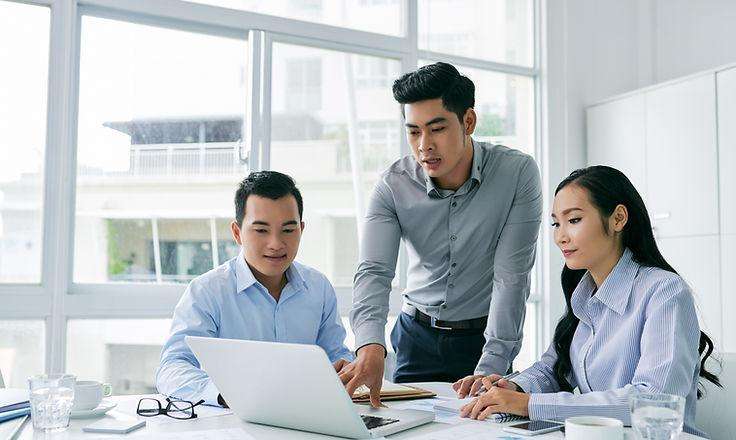 Three Asian business professionals collaborating around a laptop in a modern office, discussing digital solutions to improve business operations.